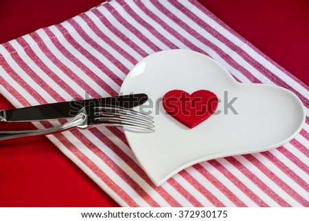 Right down on a white plate in the heart shape is a red heart next to a knife and fork on red background of the tablecloth and napkins with white stripes. Heart on a plate. Horizontal. Foto stock © 