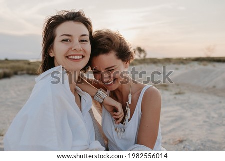 smiling two young women friends having fun on the sunset beach, queer non-binary gender identity, gay lesbian love romance, boho summer vacation style wearing jeans