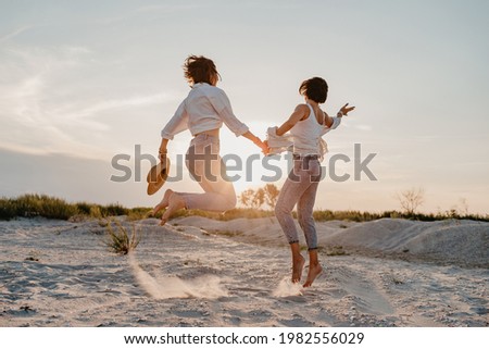 happy two young women having fun on the sunset beach, queer non-binary gender identity, gay lesbian love romance, boho summer vacation style wearing jeans