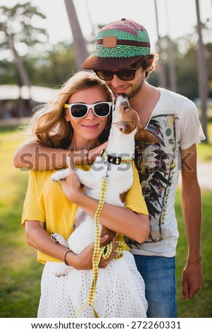 young stylish hipster couple in love holding a dog at the tropical park, smiling and having fun during their vacation, wearing sunglasses, cap, yellow and printed shirt, romance