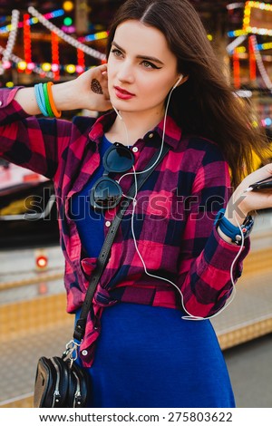 young hipster stylish beautiful woman listening music mobile phone, earphones, blue dress and checkered shirt at the amusement park, enjoying the summer day, cool accessories, mehndi drawing on hand
