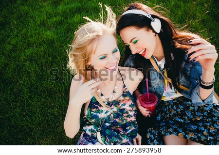 two young beautiful happy stylish hipster girls, friends together, cocktail, drink, denim outfit, smiling, fashion, cool accessories, vintage style, crazy having fun, park, sitting, grass, laughing