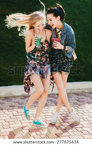 two young beautiful happy stylish hipster girls, friends together, cocktail, drink, denim outfit, smiling, happy, fashion, cool accessories, vintage style, having fun, park, crazy mood, laughing