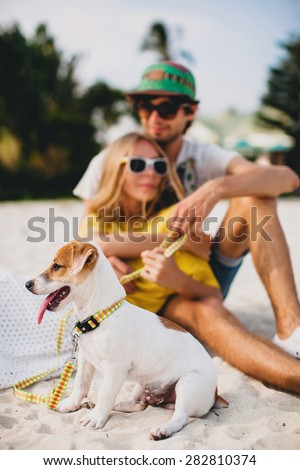 young stylish hipster couple in love playing dog puppy jack russell in tropical beach, cool outfit, romantic mood, having fun, sunny, man woman together, vacation, lying on the sand