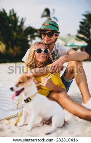 young stylish hipster couple in love playing dog puppy jack russell in tropical beach, cool outfit, romantic mood, having fun, sunny, man woman together, vacation, lying on the sand