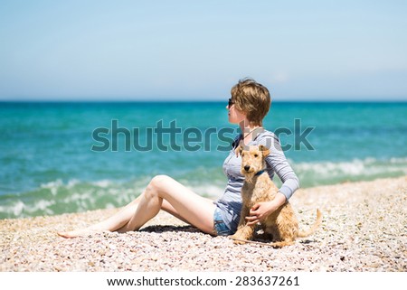 Beautiful girl with blonde hair in a blue blouse sitting on the beach with a dog