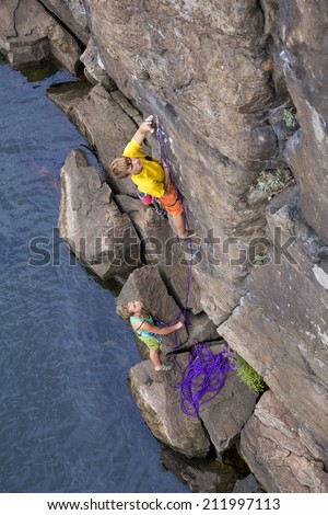 Climbing partners make ascent on to the rock wall. Two climbers - male and female - climb rocky wall over the river rocky beach. Young male climber is leading, female teenager is belaying him.
