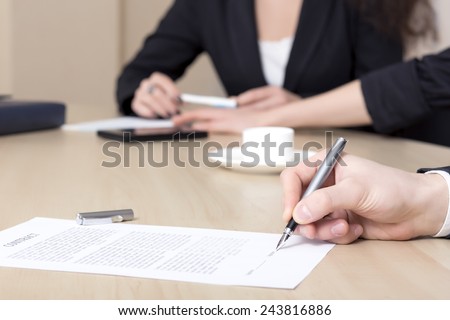 Male businessperson signs contract. Close up of female hand signing formal paper on the office table. The business counterpart on the background