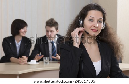 Cheerful female customer support officer. Portrait of smiling cheerful customer support phone operator in headset with her co-workers on the background