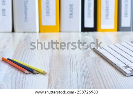 Desk of creative person.\
Side view on light wooded desk with color pencils and keyboard, with stack of folders on the background
