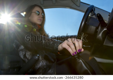 Confident female driver.\
Young woman drives car, focus on her hand switching transmission grip. Steering wheel on the right, blues sky in the window, bright sunbeams on the left