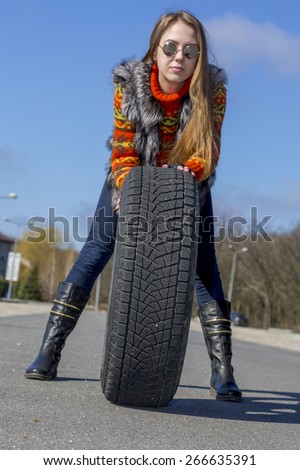 Female biker rolls big wheel.\
Young female, dressed in orange sweater and fur vest, holds big black wheel with winter tire