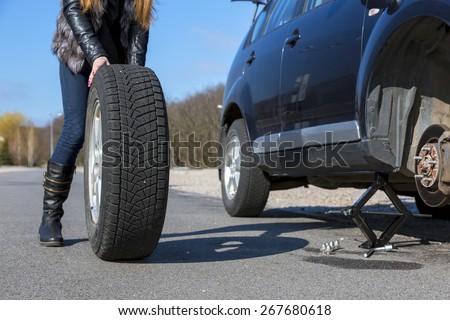 Female driver repairs car. Body of young female, dressed biker style, rolling big wheel towards broken car.