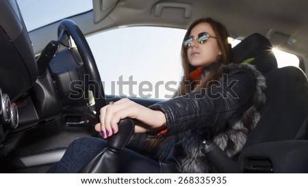 Confident female racer drives car. Young woman drives car, focus on her hand switching transmission grip Biker dress black leather jacket, sunglasses, bright sunbeams on right corner
