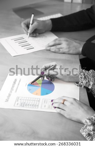 Womankind make office life brighter. Hands of male and female working on printed charts located on the office desk. Black and white background, but one chart and female nails and finger ring colored