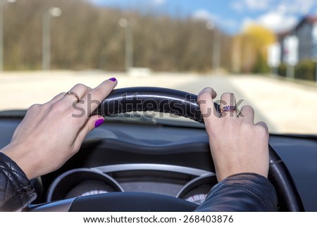 Hands of female driver on steering wheel.\
Woman holds the passenger car steering wheel, blurred road, colorful forest landscape, blue sky on the background. Hands skin is carefully smothered