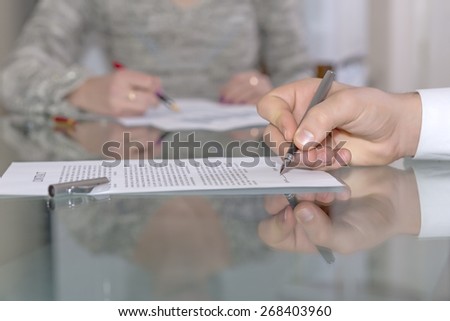 Male hand signing formal paper. Close-up hand on foreground working with paper sheet fountain pen female body on background glass office desk with reflection