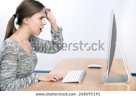 Surprising news. Young beautiful lady expresses shock looking on large computer screen, smart casual dress, beige office desk, wireless keyboard and mouse, light grey background