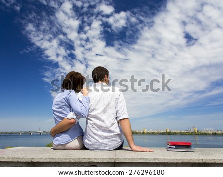 Young couple and cloudy sky. Two people male and female sitting on railing embracing in front of urban view with river and blue sky clouds
