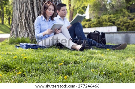 Grass and flowers and two people reading. Outdoor park lawn green fresh grass yellow dandelion large tree young man woman casual dress read tablet PC book focus on grass and flowers people blurred