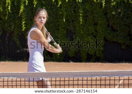 Young tennis athlete ready to return a ball.\
Elegant girl tennis white dress with skirt swinging racket to hit coming ball green fence background