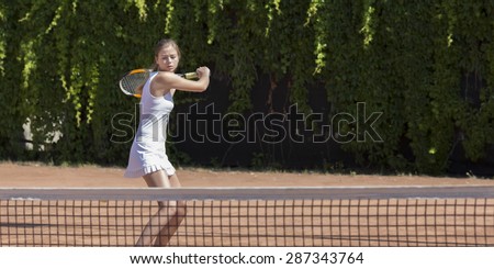Well prepared to defend. \
Young female tennis athlete swinging racket to meet coming ball white dress miniskirt outdoor play court dark green fence background orange clay ground copy space on right