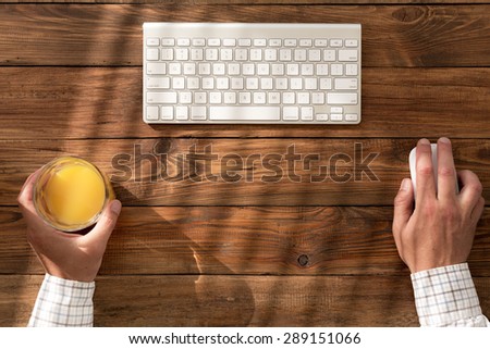 Top view of man at natural wooden desk.\
Clean working space at vintage table businessman holding orange juice glass compute mouse and sunbeams coming throw venetian blinds