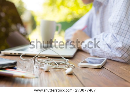 Remote office concept Business style dressed man sitting at natural country style wooden desk with electronic gadgets around working on laptop drinking coffee sunlight and green terrace on background