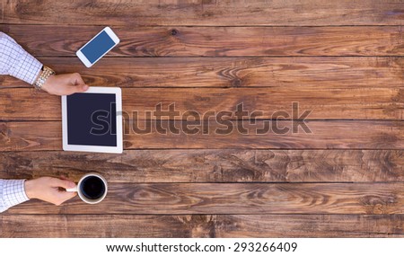 Natural wood desk directly top view man arms holding white tablet PC coffee mug black screen cell phone casual dress shirt cuffs red light brown board plank browsing internet