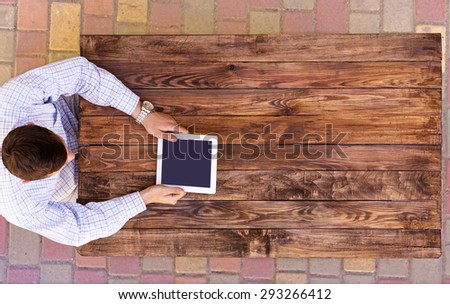From above image man browsing gadget at empty wood desk.\
Natural wood desk directly top view man arms holding white tablet PC black screen casual dress shirt light brown board plank browsing internet