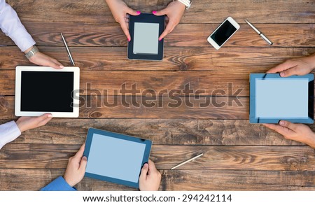 Group of people with varied gadgets at wooden desk. Direct from above view on hand of people holding tablets and electronic books under natural planked wooden desk.