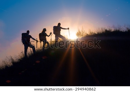 Three people family silhouettes on vacation.\
Group backpackers moving up toward grassy veld hill uprising colorful sun and rainbow clouds on background