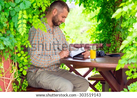 Travel diary. Bearded man sitting at vintage design wooden arbor and writing notes into his notepad having glass of orange juice and photo camera on table surrounded green flora sun light background