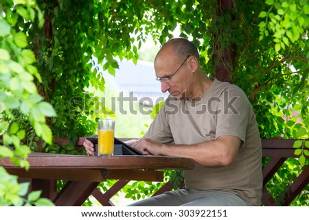 Casual dressed man with gadget and glass of orange juice sitting in wood arbor.\
Aged man pointing into screen of black tablet PC sitting inside wooden pavilion in green park with curled tree leafs