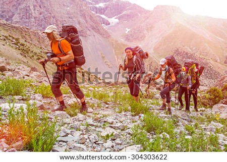 Extreme climbers scrambling up.\
Group people approach high altitude mountain climbing camp with heavy backpacks tons alpine gear walking on rocky path trail at peaks glaciers snow sun sky background