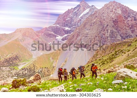 People traveling in mountains.
Large group of tourists of different sex ethnic nation race age young and old man woman walking up on rocky path with green grass forest and mountain peaks around