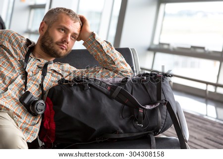 Traveler looking into far.\
Portrait of bearded man inside airport terminal pensive casual shirt and travel pants sitting next to his backpack luggage large bright window on background