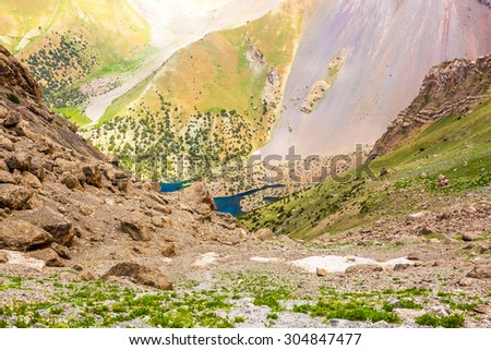 Mountain lake view in Fan Mountains of Tajikistan. From above perspective on colorful terrain with green grass brown and red rocks some forest and deep blue lake in the middle with some pieces of snow