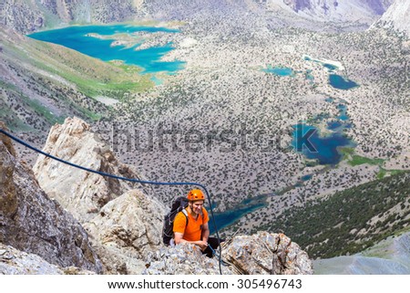 Mountain climber over abyss. Elder man orange jacket protection helmet holding belaying rope rocky cliff arranging descent wild vivid color mountain lakes on background