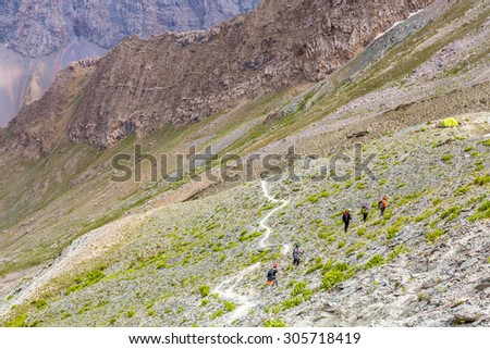 White mountain footpath and group of hikers.\
Mountain landscape with green grass and red orange rocky peaks white dusty trail and group of travelers walking toward camping tents