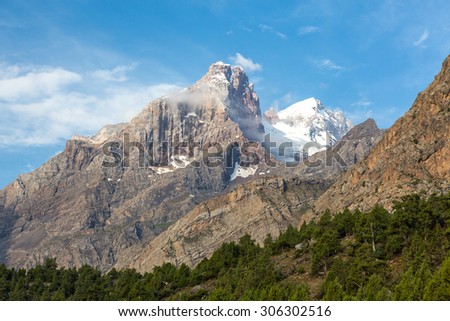 Mountain peak in Fan Mountains.\
Landscape with layer of green forest at bottom orange tone rocky peak with snow and glaciers on top on blue sky