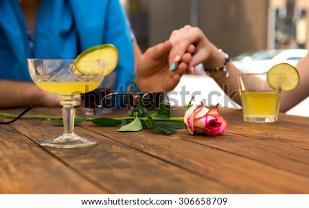 Romantic scene flower on foreground Vintage natural wood cafe table with beverage glasses rose flower on foreground  lovers holding hands of each other young male female vacation style environment