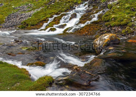 Mountain stream and meadow.\
Flowing mountain stream of river with several creeks effluent from out of ground grassy meadow along shore orange red color stones cleaving water