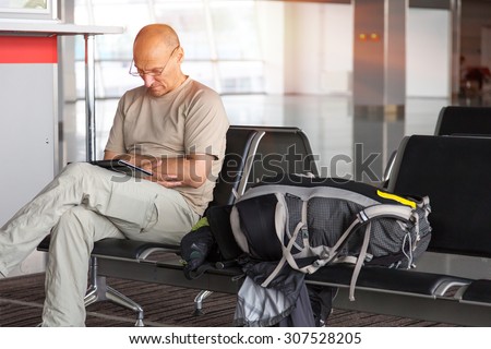Waiting for boarding.\
Mature traveler sitting business class lounge airport terminal carry on luggage backpack using electronic device connect internet t-shirt travel pants sun back light background
