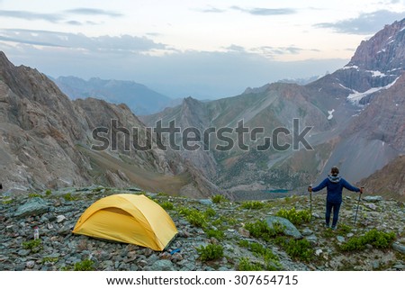 Hiker with walking poles and climbing tent.\
Man observing scenic evening view of mountain valley yellow camping tent on grassy rocky place
