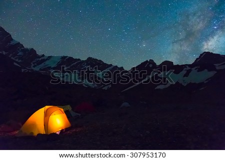Night mountain landscape with illuminated tent.
Silhouettes of snowy mountain peaks and edges night sky with many stars and milky way on background illuminated orange tent on foreground