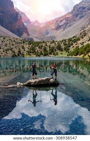 Pair of Female Hikers in the middle of Lake Energetic Bodies of Active Lifestyle People Staying on Stone Among Vivid Colored Mountain Lake Shining Sun and Sunbeams Coming Throw Peak Range Background