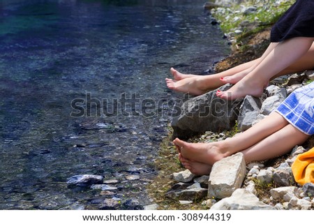 Mountain lake and female feet.\
Three pairs of female legs relaxing on the waterfront of pristine lake focus on the middle feet with red nail pedicure