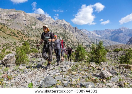 People traveling in mountains.\
Large group of tourists of different sex ethnic nation race age young and old man woman walking up on rocky path with green grass forest and mountain peaks around