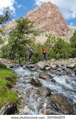 Two hikers crossing fast flowing river.\
People going across mountain creek with fast streaming water jumping on the rocks green meadow and forest along river mountain landscape and sky on background